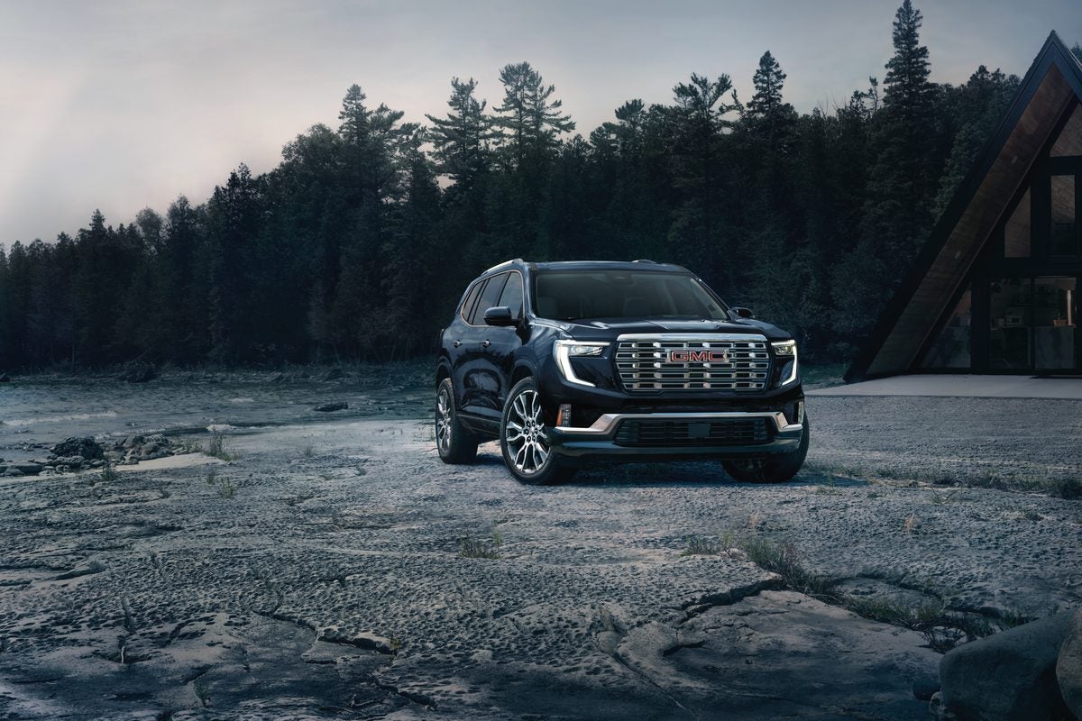 Black 2027 GMC Acadia Denali on a rocky lakeshore with pines and an A-frame behind