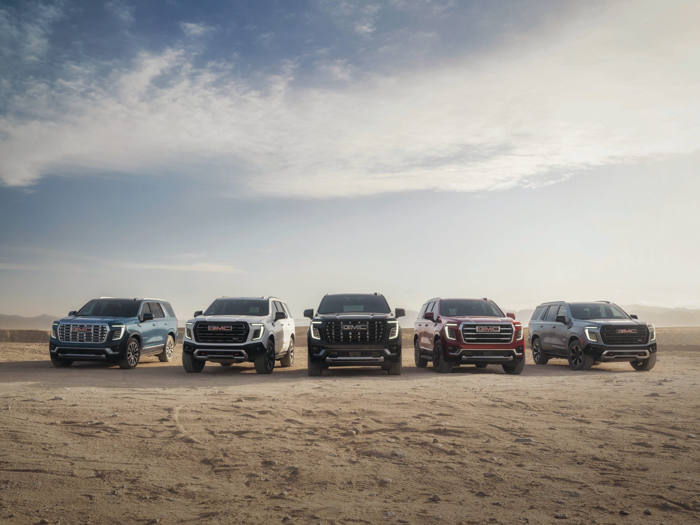 Five GMC Yukon and Acadia SUVs lined up on open sand, GMC's 2027 SUVs photographed together