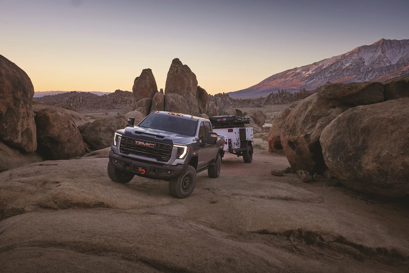 2027 GMC Sierra HD pickup with a small camping trailer behind it, parked on desert rocks at dusk with a snow-capped ridge in the distance