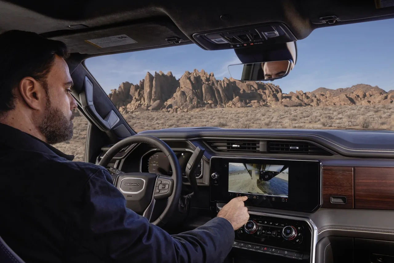 Driver reaching for the center touchscreen of a 2027 GMC Sierra HD, which is showing a trailer camera view
